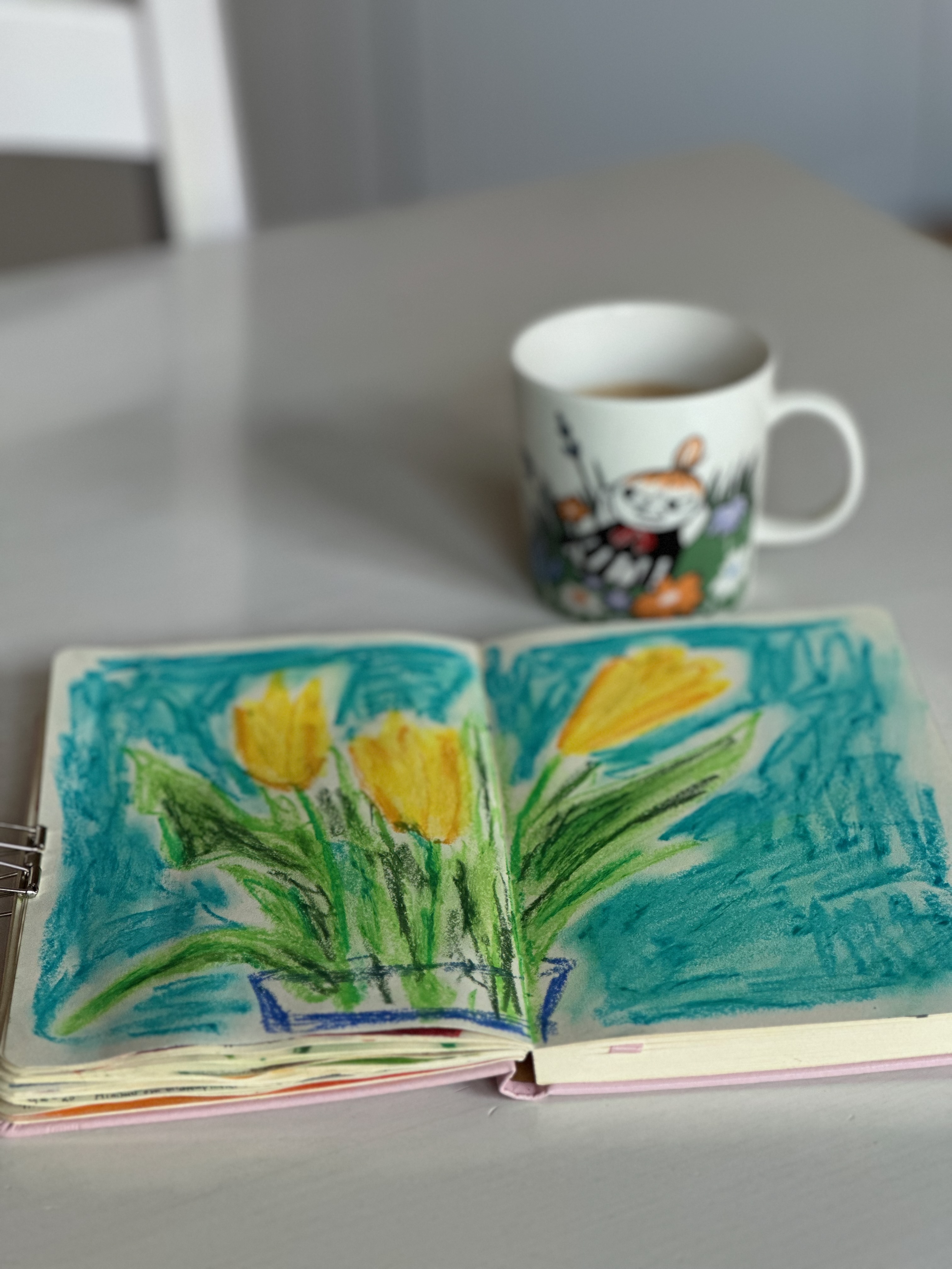 En skissbok och en kopp kaffe på ett köksbord. Skissboken är uppslagen och visar en teckning av 3 gula tulpaner på blå bakgrund. 