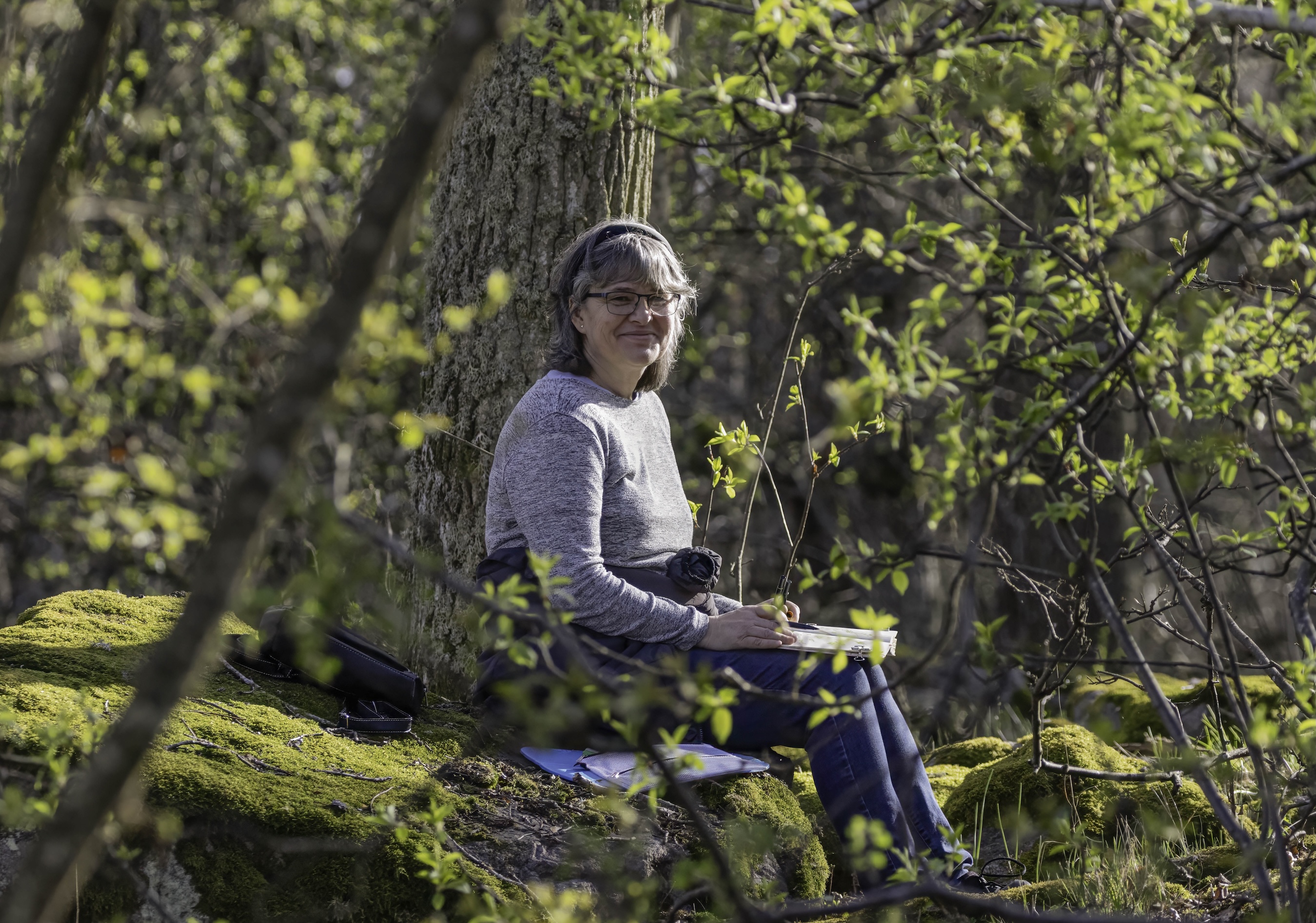Jag sitter på en mossklädd sten under ett träd och ritar i mitt skissblock, omgiven av spirande vårgrönska – en stilla stund i skogsdungen.